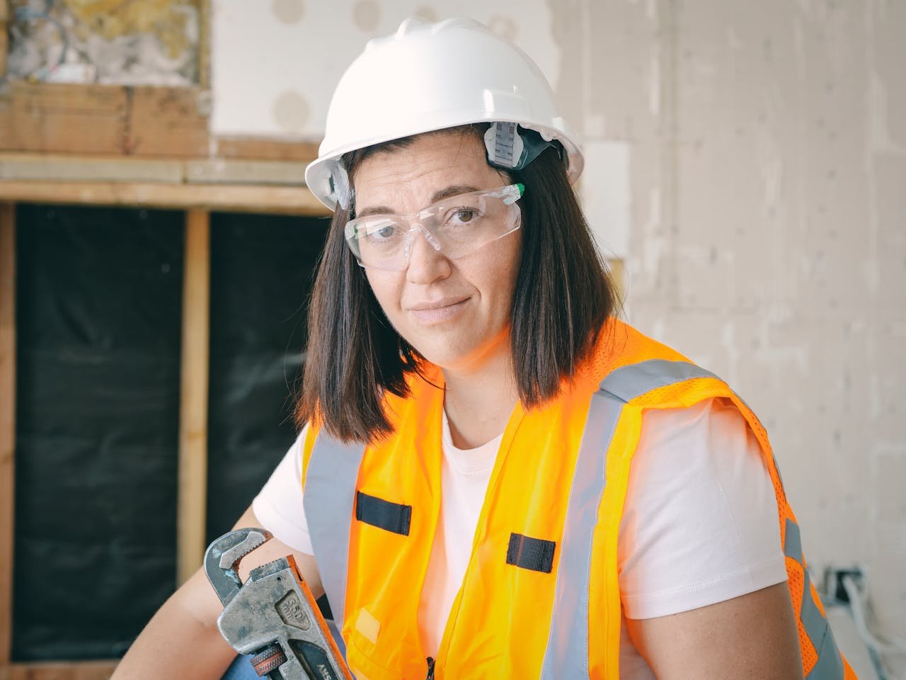 A female construction worker wearing safety gear holding a wrench indoors.